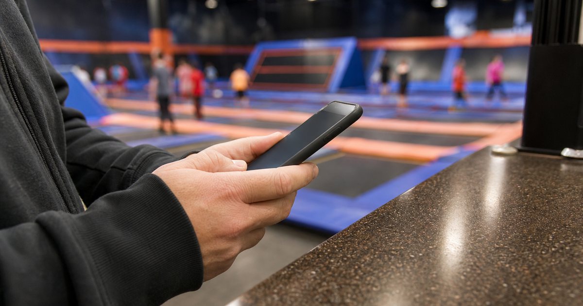 Close-up of a person's hands holding a smartphone at the reception counter of a trampoline park, colorful padded arena blurred in the background, the screen angled away from the camera showing no readable content
